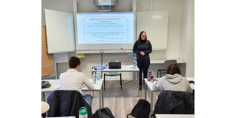 Dozentin steht vor der Klasse und unterrichtet; Studierende sitzen an Einzeltischen.
