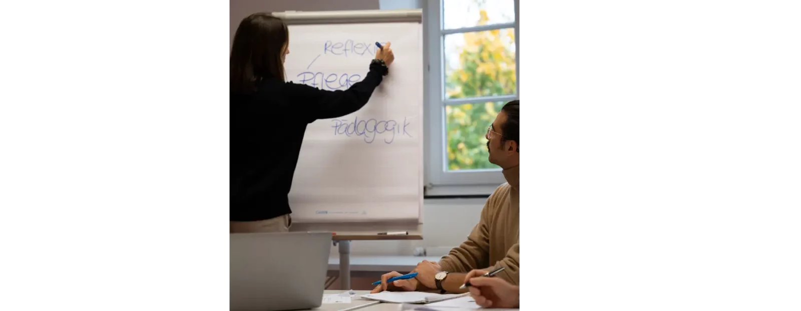 Eine Frau schreibt auf ein Flipchart in einem Seminarraum. Eine weitere Person schaut zu. Auf dem Flipchart stehen die Worte „Pflege“, „Pädagogik“ und „Reflexion“.