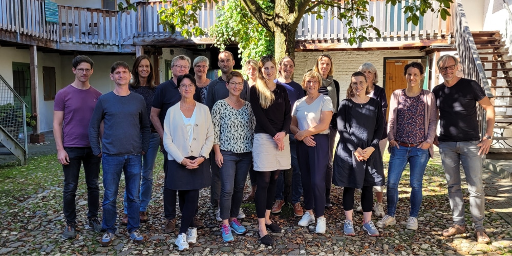 Eine Gruppe von 17 Erwachsenen steht unter einem großen Baum in einem gepflasterten Innenhof für ein Gruppenbild.