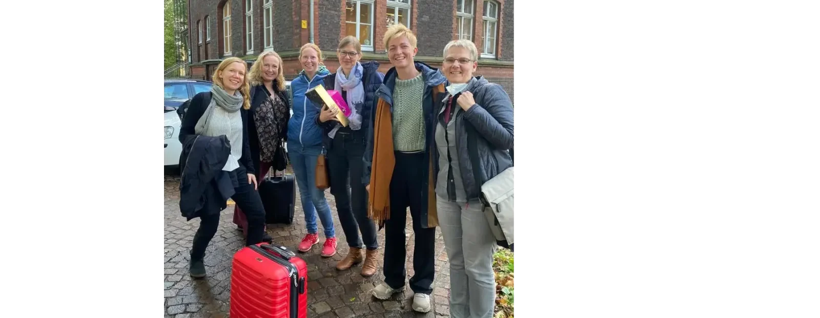 Sieben Personen stehen lachend mit Koffern und Taschen vor einem Backsteingebäude, vermutlich nach oder vor einer Reise.
