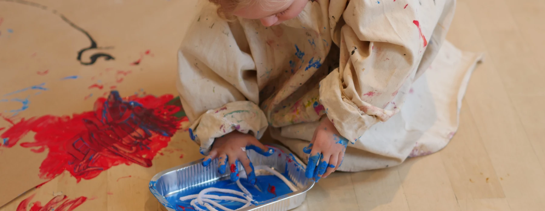 Ein Kind in einem Malerkittel kniet auf dem Boden und malt mit den Händen in blauer Farbe auf Papier