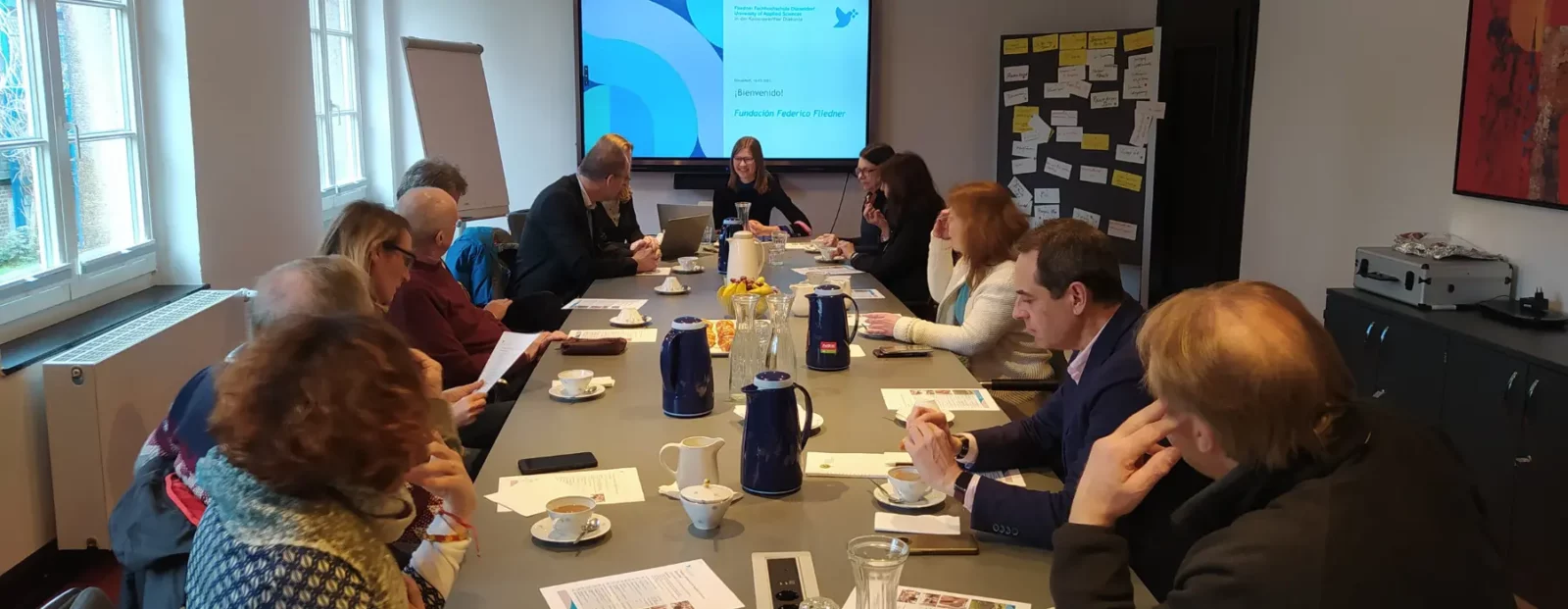 Mehrere Personen sitzen um einen langen Tisch mit Getränken, Tassen und Papieren. Eine Präsentation mit dem Titel „¡Bienvenido! Fundación Federico Fliedner“ ist auf dem Bildschirm zu sehen.