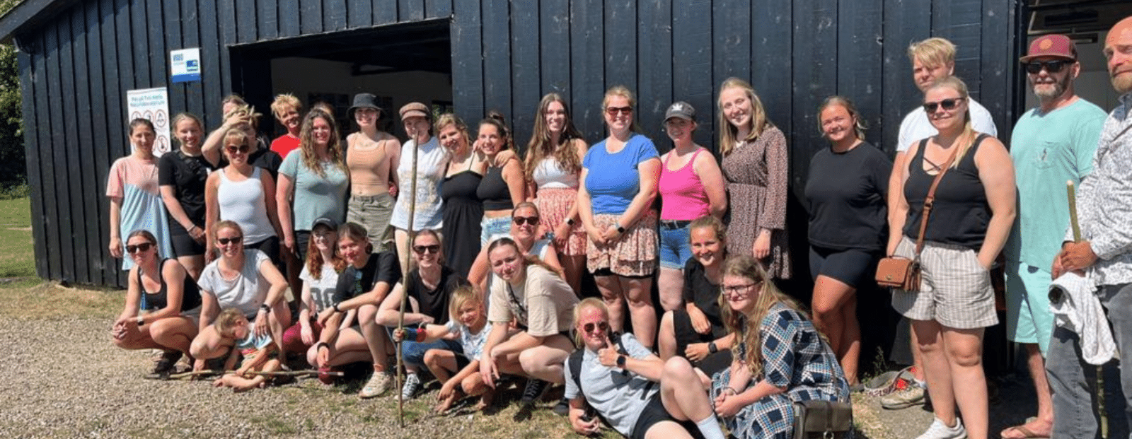 Große Gruppe aus etwa 30 Personen, darunter Kinder und Erwachsene, steht oder hockt vor einem dunklen Holzgebäude in sommerlicher Kleidung. Die Stimmung ist entspannt und freundlich.
