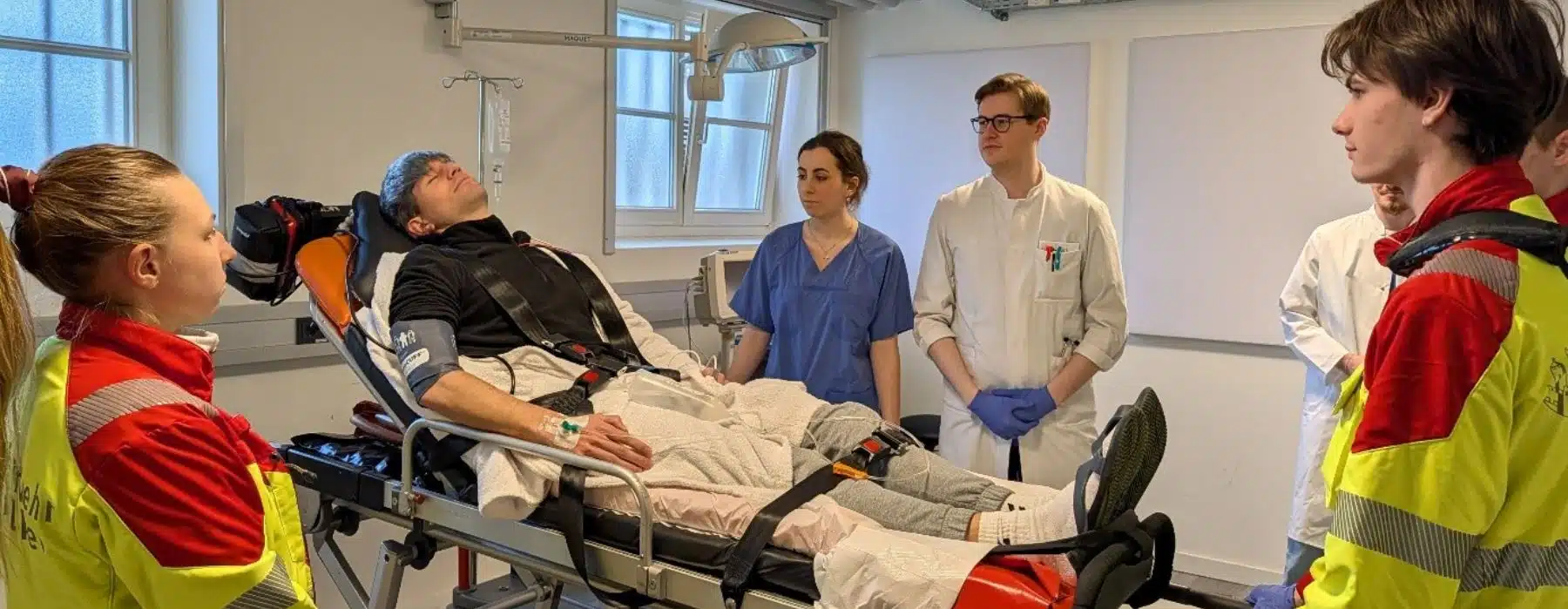 Im SkillsLab der Fliedner Fachhochschule wird ein Notfall simuliert. Auf der Trage liegt ein Auszubildender, im Vordergrund stehen zwei junge Sanitäter:innen und neben der Trage steht eine Pflegekraft und ein Arzt