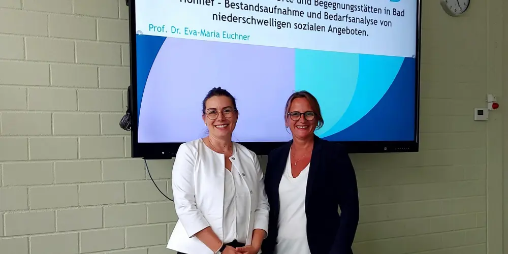 Prof. Dr. Eva-Maria Euchner steht mit einer anderen Frau vor einem Bildschirm, auf dem geschrieben steht: Abschlussbericht: Dritte Orte und Begegnungsstätten in Bad Honnef - Bestandsaufnahme und Bedarfsanalyse von niederschwelligen sozialen Angeboten